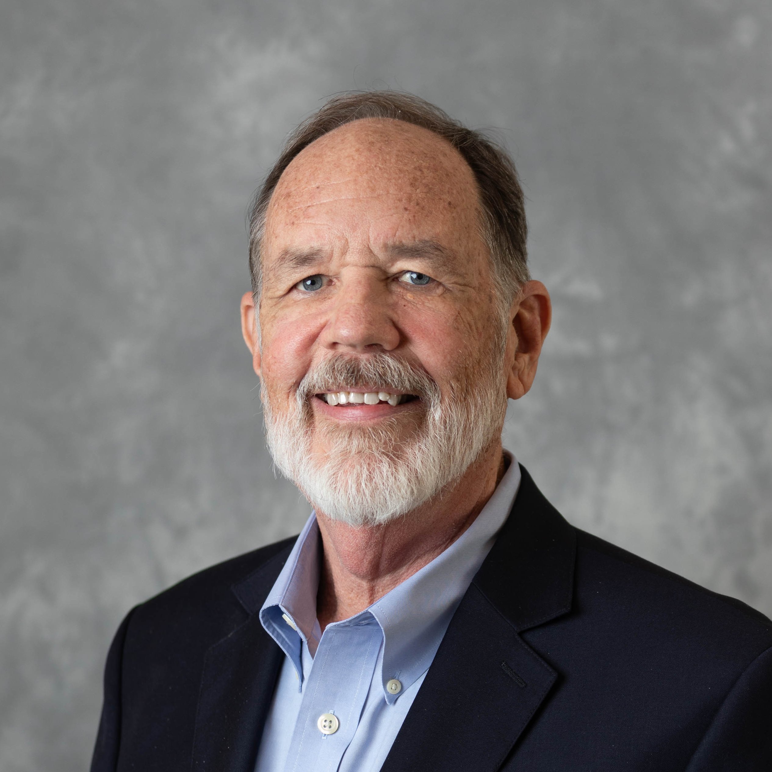 Professional headshot of a smiling man with a beard, wearing a navy blazer and light blue shirt, against a textured gray background, representing expertise in copywriting services.